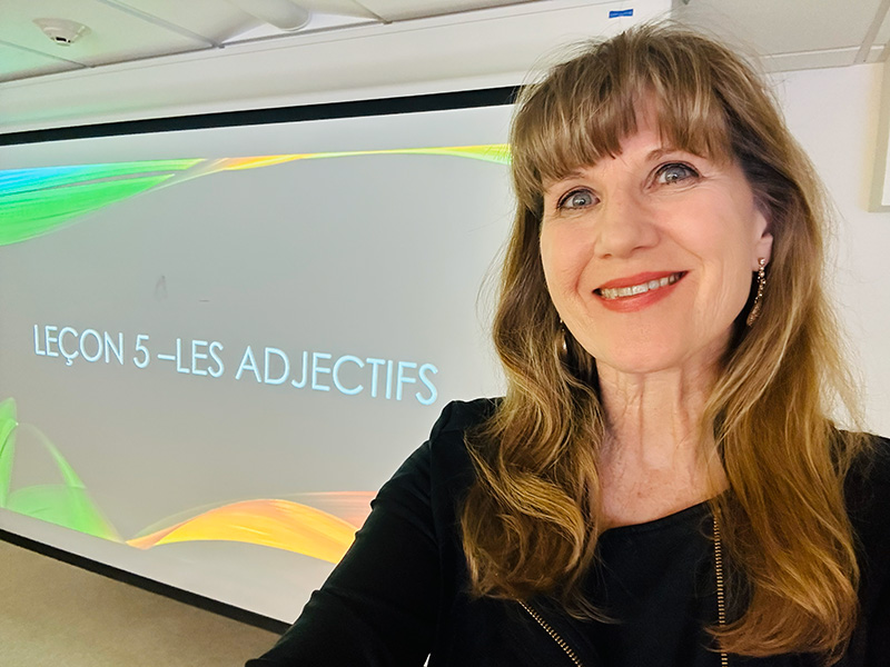 A female instructor with long blond hair is smiling and standing in front of a large screen that displays the French words Lecon 5 - Les Adjectifs.