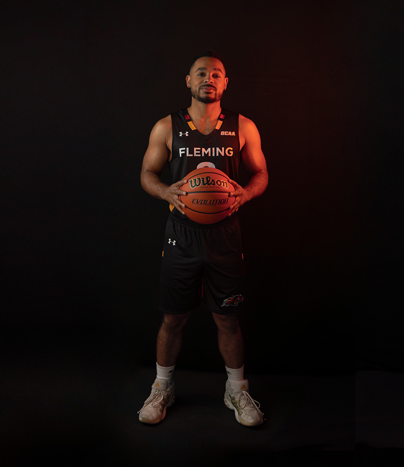 A male student wearing a Fleming Phoenix basketball uniform stands intently holding a basketball in front of him.