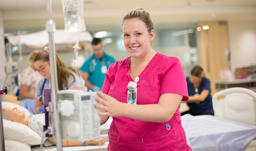 A female Fleming College practical nursing student in a pink uniforn is shown operating medical equipment in a clinical lab. There is are male and female nursing students training in the background.