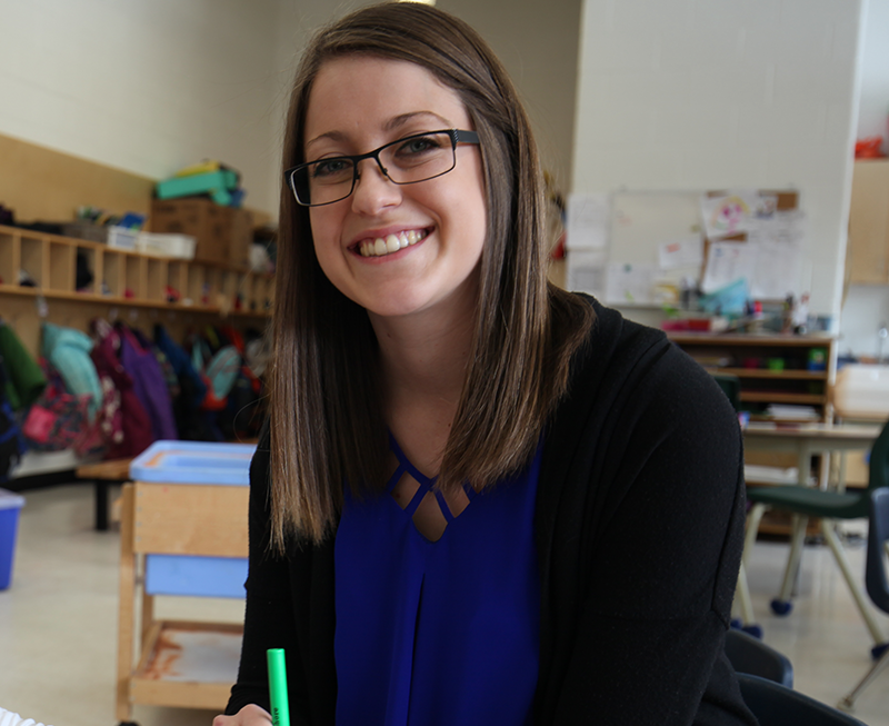 A female Early Childhood Educator in a classroom setting wears glasses and is smiling.