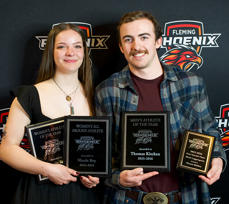 A male and female stand together smiling and holding awards.