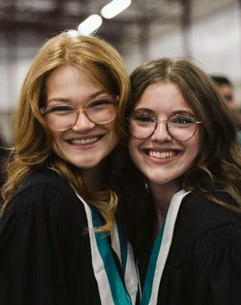 Two female graduates stand together with huge smiles wearing black gowns and teal and white graduate collars.
