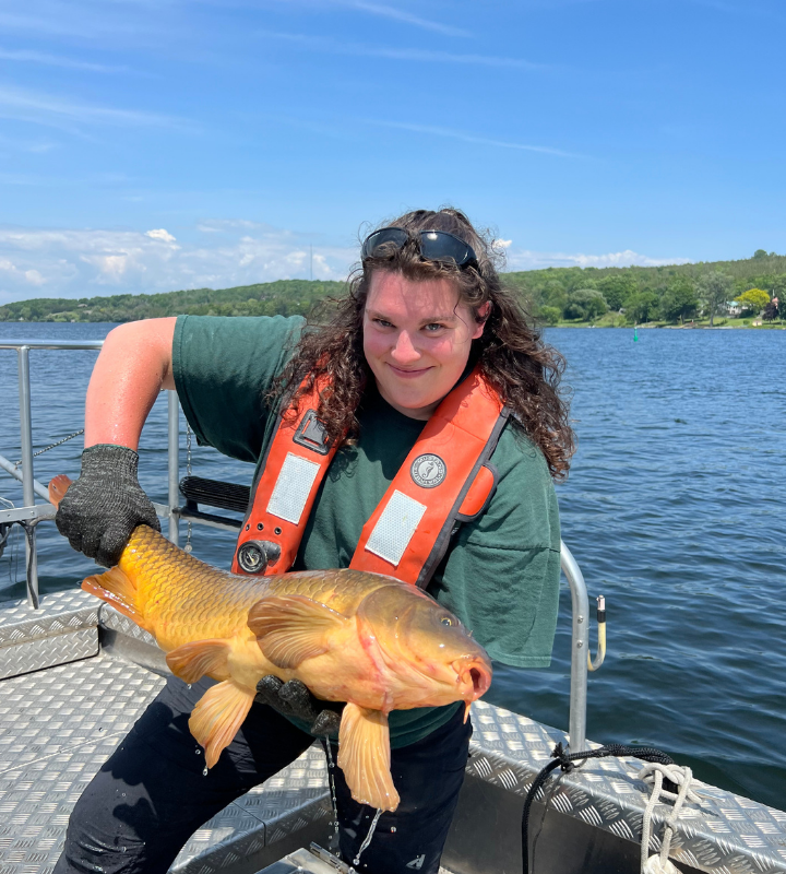 A woman wearing an orange lifejacket stands in a fishing boat smiling and holding a large fish with both hands.