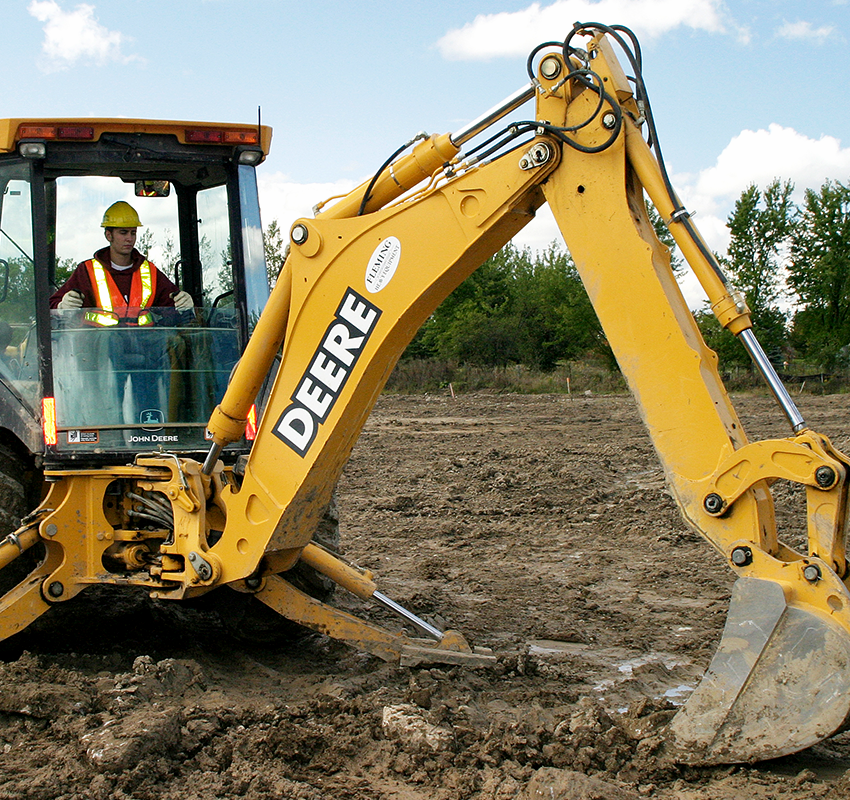 A male fleming college students operating a large yellow excavator in a dirt covered landscape.