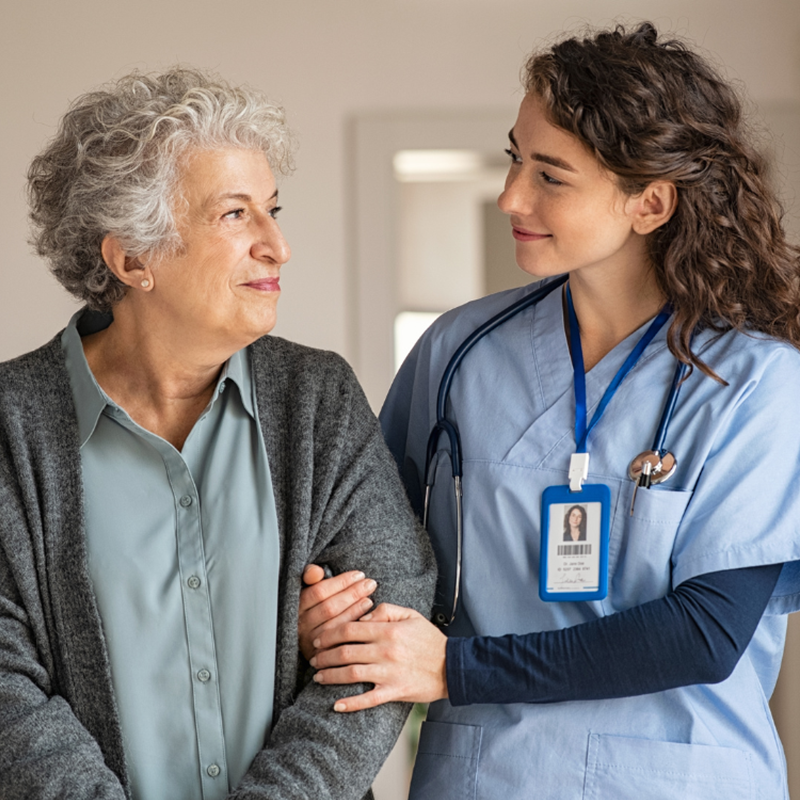 A personal support worker in a blue uniform stands with her arm around another senior women's arm in support. They are looking at each other and smiling.