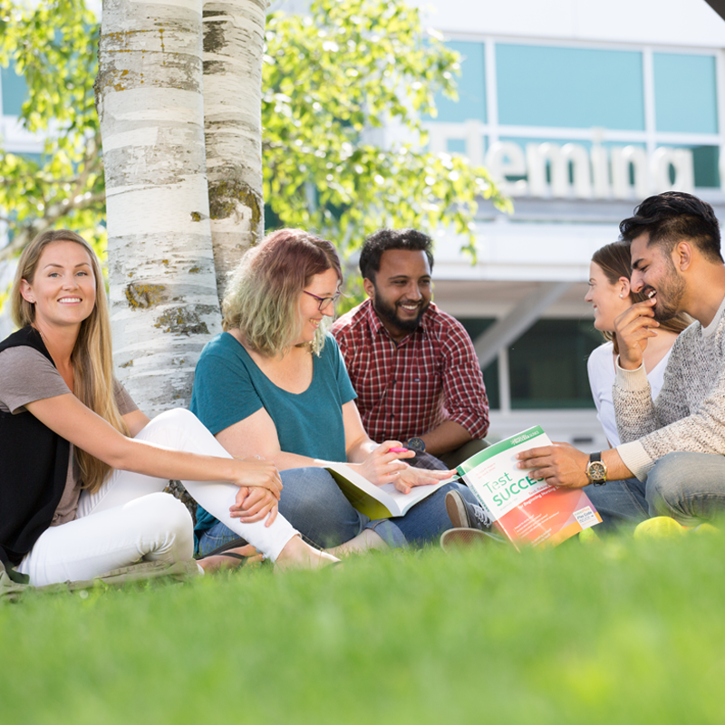 A group of male and female students smiling and looking at a text book. They are sitting in front of the Fleming College entrance.