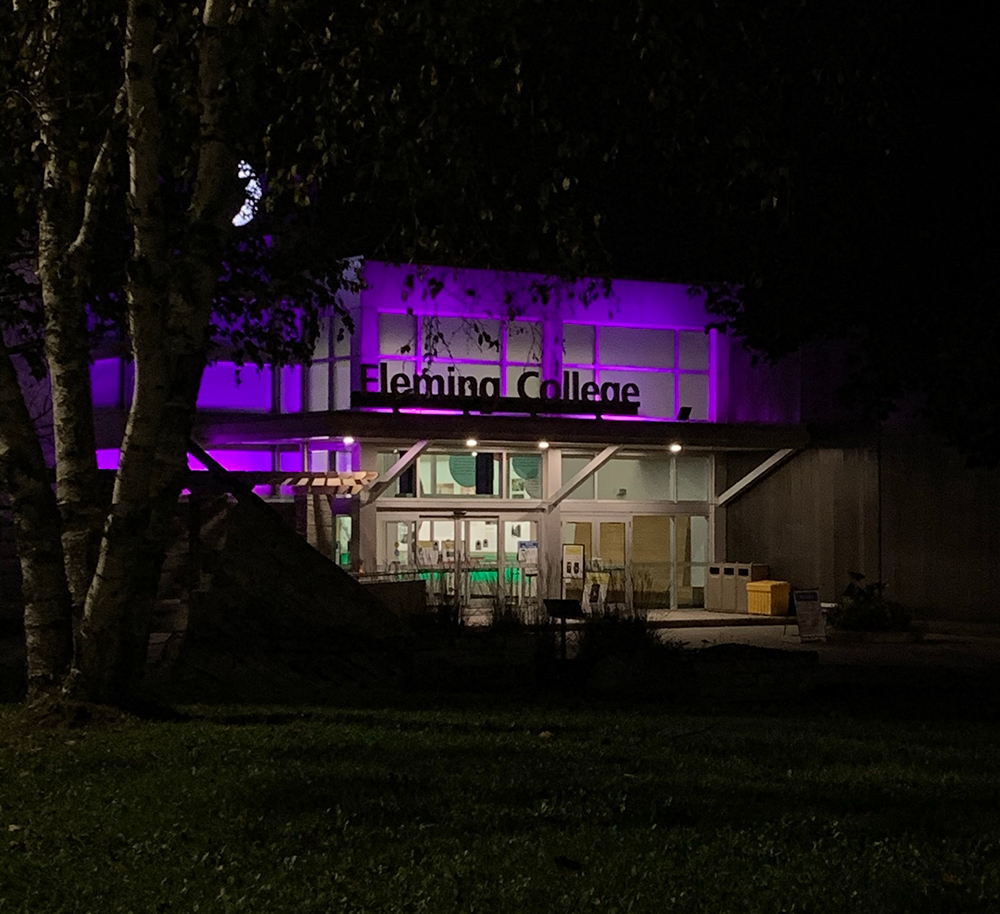 Surrounded in darkness, the front entrance of Fleming College is lit up in neon purple and blue lights.