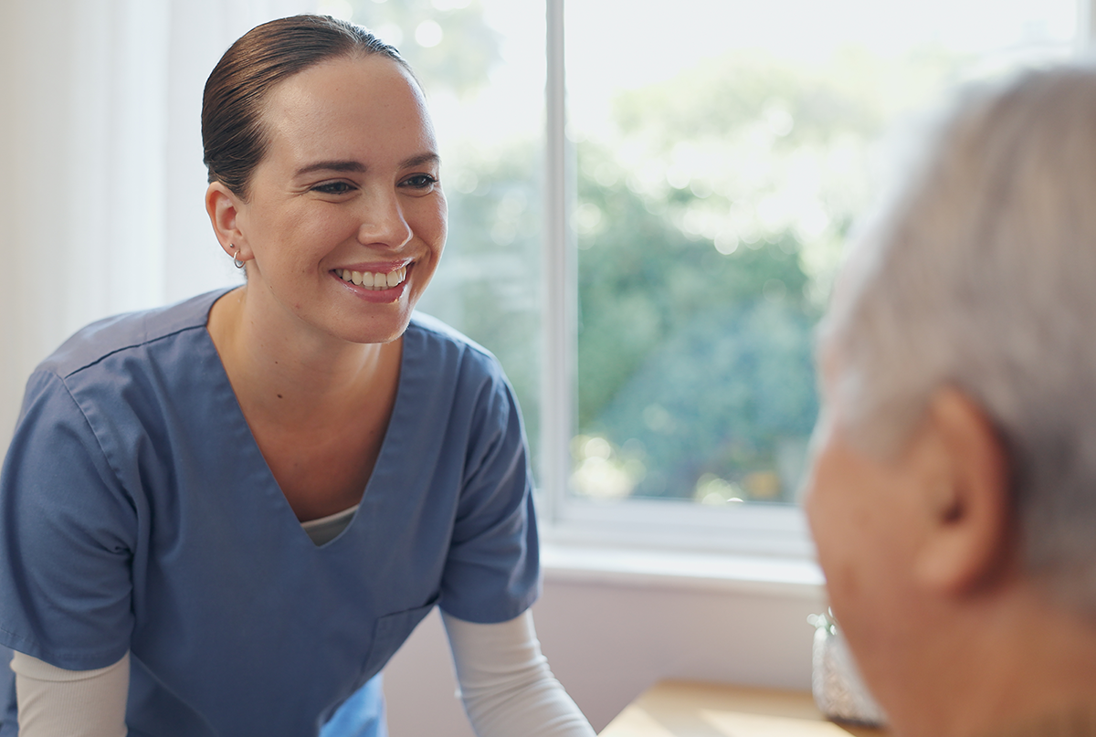 A women in a blue nurses uniform smiles while looking at a elderly patient.