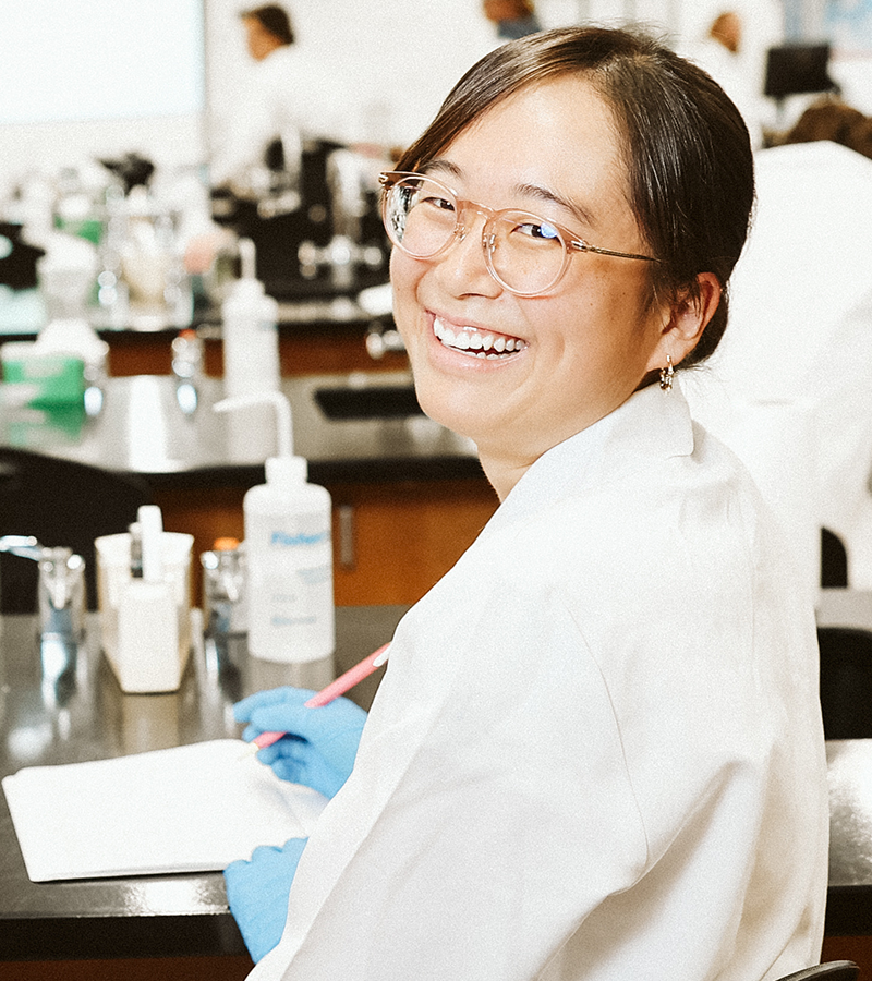 A young woman seated at a medical lab station is turned back and smiling.