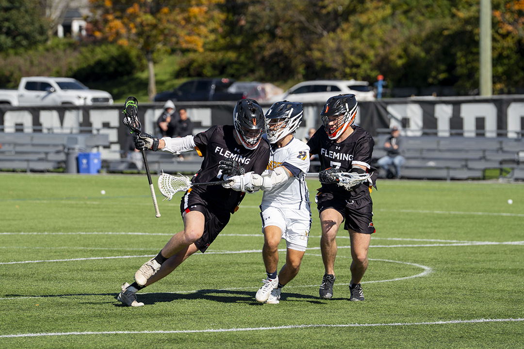 Two Fleming Phoenix lacrosse players on a sports field in physical contact of a competing player who is trying to get the lacrosse ball.