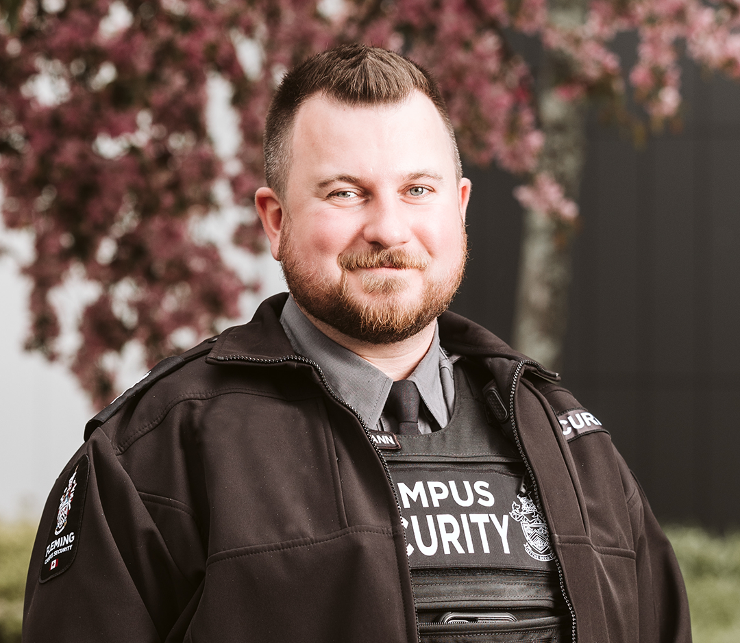 A man dressed in a security guard uniform stands in front of a maple tree smiling.